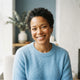 A smiling woman with short, dark curly hair, wearing a soft sky blue sweater, against a slate blue and bright white backdrop.